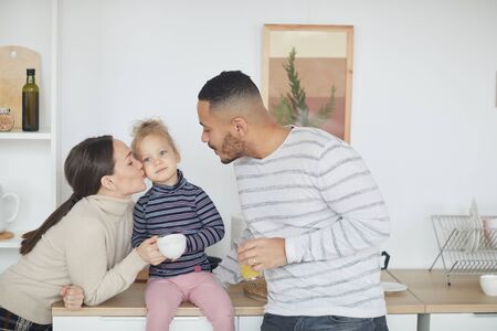 Portrait of happy mixed-race family kissing cute little girl while enjoying breakfast together in modern kitchen interior, copy spaceの写真素材
