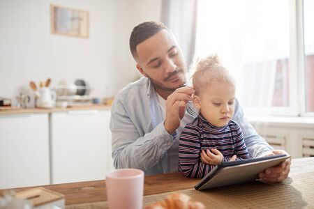 Warm-toned portrait of caring father with cute little girl using digital tablet together while sitting at wooden table in cozy kitchen interior, copy spaceの写真素材