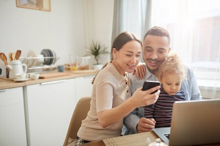 Warm-toned portrait of modern mixed race-family using computer devices while sitting in cozy kitchen interior with cute little daughter, copy spaceの写真素材