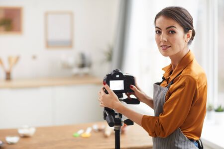 Waist up portrait of beautiful woman setting up camera and smiling while filming cooking tutorial in...