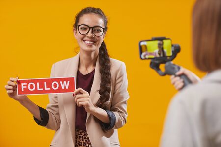 Waist up portrait of contemporary young woman holding Follow word while filming video for beauty and lifestyle channel standing against yellow background, copy spaceの写真素材