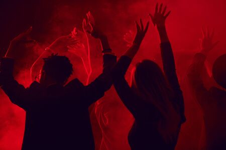Dark silhouettes of young people dancing and raising hands while enjoying party in smoky nightclub lit by red light, copy spaceの写真素材