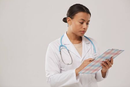 Waist up portrait of young mixed race nurse or female doctor holding clipboard while standing against white background, copy spaceの写真素材