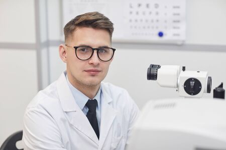 Head and shoulders portrait of male optometrist smiling at camera while posing at workplace by optic equipment, copy spaceの写真素材