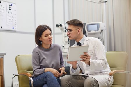 Portrait of young ophthalmologist using digital tablet while consulting female patient during eyesight check up in modern clinic, copy spaceの写真素材