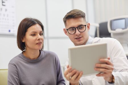 Portrait of young ophthalmologist using digital tablet while consulting female patient during eyesight check up in modern clinicの写真素材