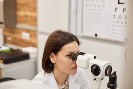 High angle portrait of young female ophthalmologist using refractometer machine during eye test in...