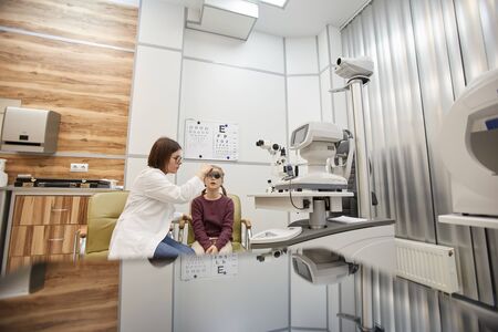 Wide angle portrait of cute teenage girl wearing trial frame during childs vision test in modern ophthalmology clinic, copy spaceの写真素材