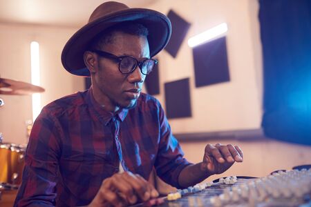 Portrait of contemporary African-American man writing music in recording studio using sound...