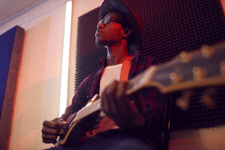 Low angle portrait of contemporary African-American man playing guitar sitting on high chair while...