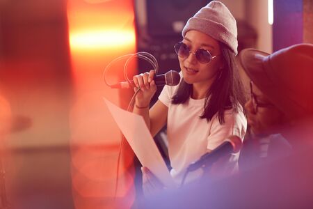 Portrait of contemporary Asian woman singing to microphone and holding lyrics sheet during rehearsal...