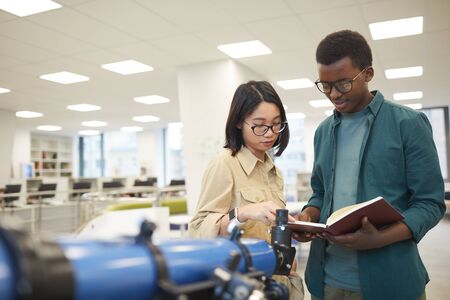 Waist up portrait of two international students reading book while standing by telescope in class or...