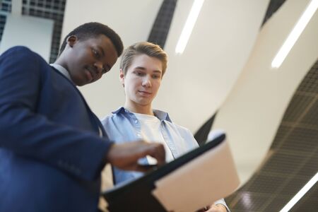 Low angle portrait of two young businessmen discussing work project while standing in office, copy...