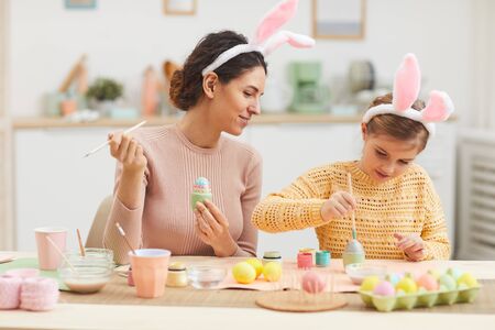 Portrait of young mother with little girl having fun while painting Easter eggs in cozy kitchen interior, both wearing bunny ears, copy spaceの写真素材