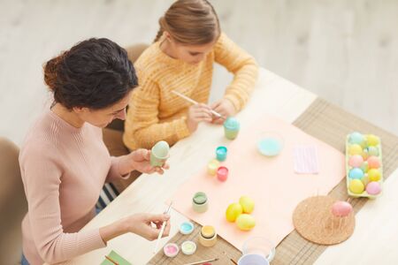 High angle view at mother and daughter painting Easter eggs pastel colors sitting at table in cozy...