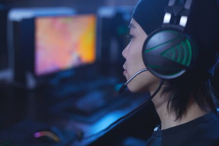 Side view close up of young Asian man playing videogames and wearing headphones in dark cyber like interior, copy spaceの写真素材