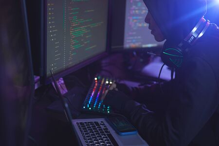Side view at unrecognizable cyber security hacker wearing hood while working on programming in dark...