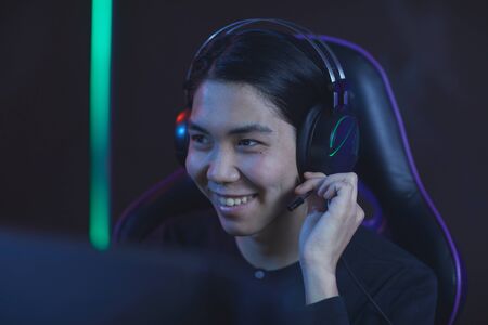 Head and shoulders portrait of smiling Asian man speaking to headset while enjoying videogames in dark room, copy spaceの写真素材