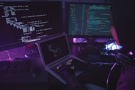 Unrecognizable young man surrounded by multiple screens programming or hacking security in dark...