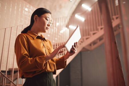 Side view portrait of young Asian businesswoman using tablet while standing by stairs outdoors, copy spaceの写真素材