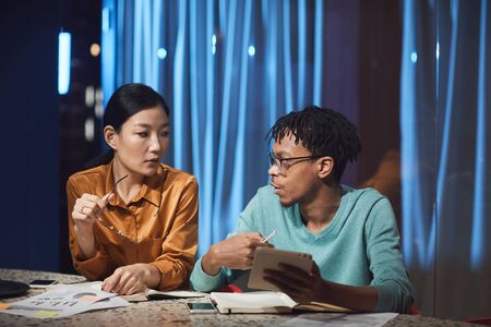 Portrait of two ethnic business people discussing project while working late in dark office, copy...
