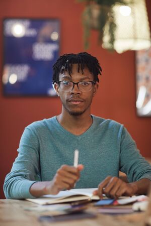 Vertical portrait of young African-American man looking at camera while sitting at table against red...