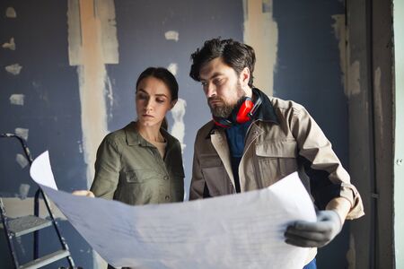 Portrait of young woman looking at floor plans with building contractor while standing against dry wall in under construction house, copy spaceの写真素材