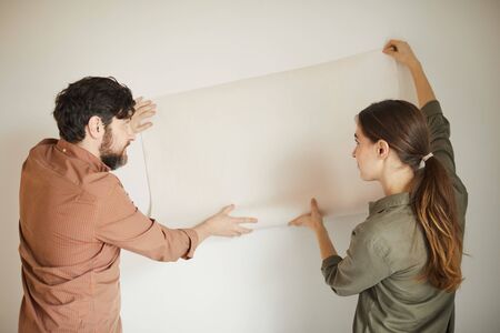 Back view portrait of married couple holding wallpaper to wall while redecorating house, copy spaceの写真素材