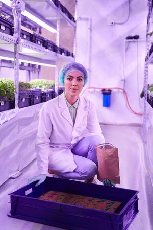 Full length portrait of female worker looking at camera while potting plants in nursery greenhouse,...