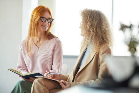 Portrait of red haired business manager talking to young intern in office and smiling happily listening to ideas, copy spaceの写真素材