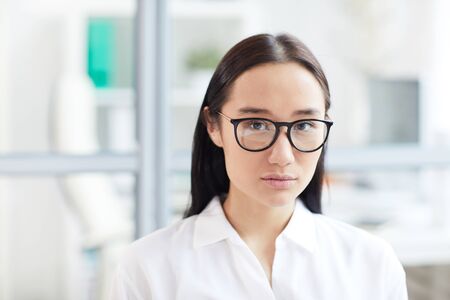 Head and shoulders portrait of young Asian businesswoman wearing glasses and looking at camera while posing in modern white office, copy spaceの写真素材