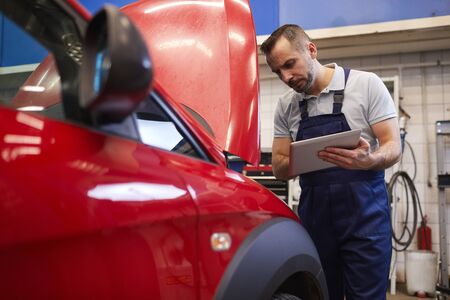 Waist up portrait of bearded car mechanic using digital tablet and looking under hood during vehicle inspection in garage shop, copy spaceの写真素材