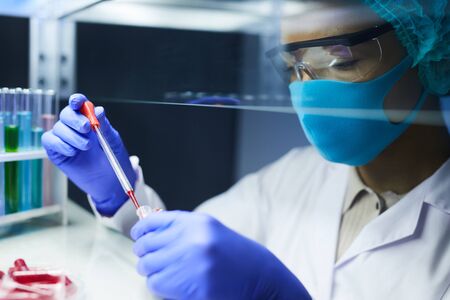 Side view portrait of female scientist wearing mask and protective gear dropping blood samples in...