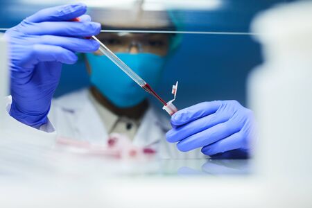 Close up of female scientist wearing mask and protective gear dropping blood samples in test tubes while working on research in laboratory, copy spaceの写真素材