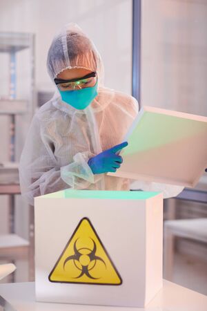 Waist up portrait of male scientist wearing biohazard gear opening box with danger sign while working on research in bio laboratory, copy spaceの写真素材