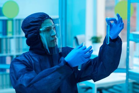 Waist up portrait of male scientist wearing biohazard gear holding test tubes while working on...