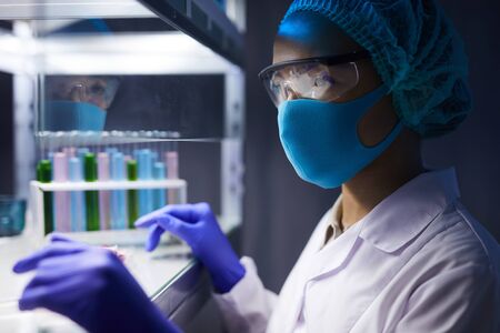 Side view portrait of female scientist wearing face mask and protective gear working on research in...
