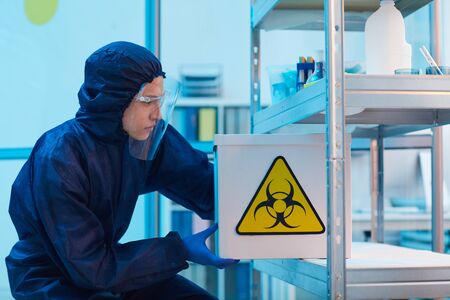 Side view portrait of male scientist wearing biohazard gear holding box with danger sign while working on research in bio laboratory, copy spaceの写真素材