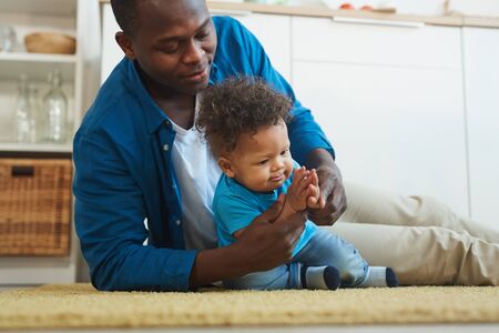 Portrait of happy African-American father playing with little boy while sitting on floor at home,...
