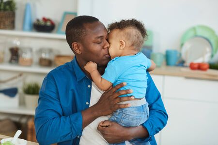 Portrait of happy African-American dad kissing little boy white sitting at kitchen table, copy spaceの写真素材