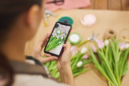 High angle close up of young woman taking smartphone photo of tulip flowers while working in flower...