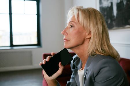Side view at pensive mature businesswoman holding smartphone wile waiting in cafe or office, copy...