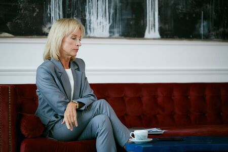 Side view at modern mature businesswoman looking away pensively while sitting on velvet sofa in cafe, copy spaceの写真素材