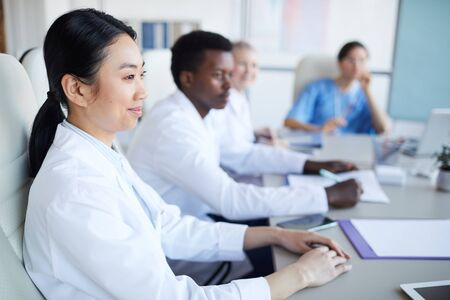 Side view at multi-ethnic group of doctors sitting at table during medical conference, focus on smiling Asian woman in foreground, copy spaceの写真素材