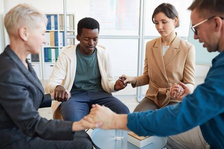 Multi-ethnic group of people holding hands in prayer while sitting in circle in support group, copy spaceの写真素材
