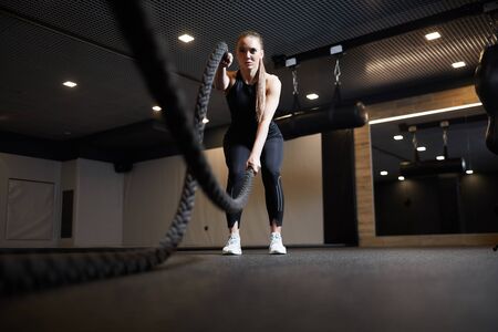 Full length wide angle portrait of focused young woman doing battle rope exercises during cross...