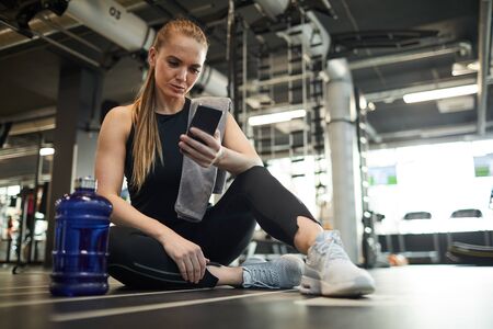 Full length portrait of modern young woman using smartphone while sitting on floor in gym and taking...