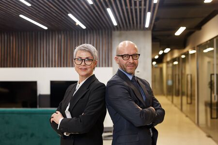 Waist up portrait of two successful mature business people standing back to back with arms crossed...