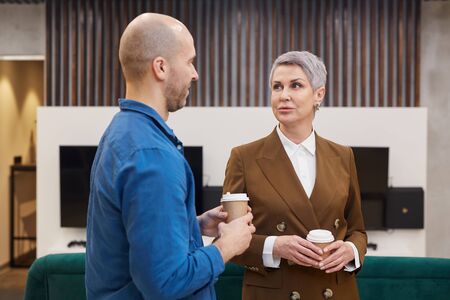 Waist up portrait of modern mature businesswoman talking to colleague and holding coffee cup during...