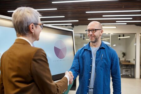 Waist up portrait of bald adult man shaking hands with businesswoman while standing against...